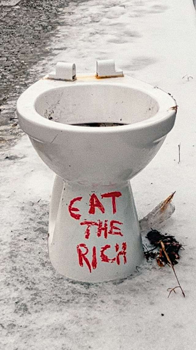 A toilett without seat standing on the sidewalk with the words "EAT THE RICH" written on it in red.